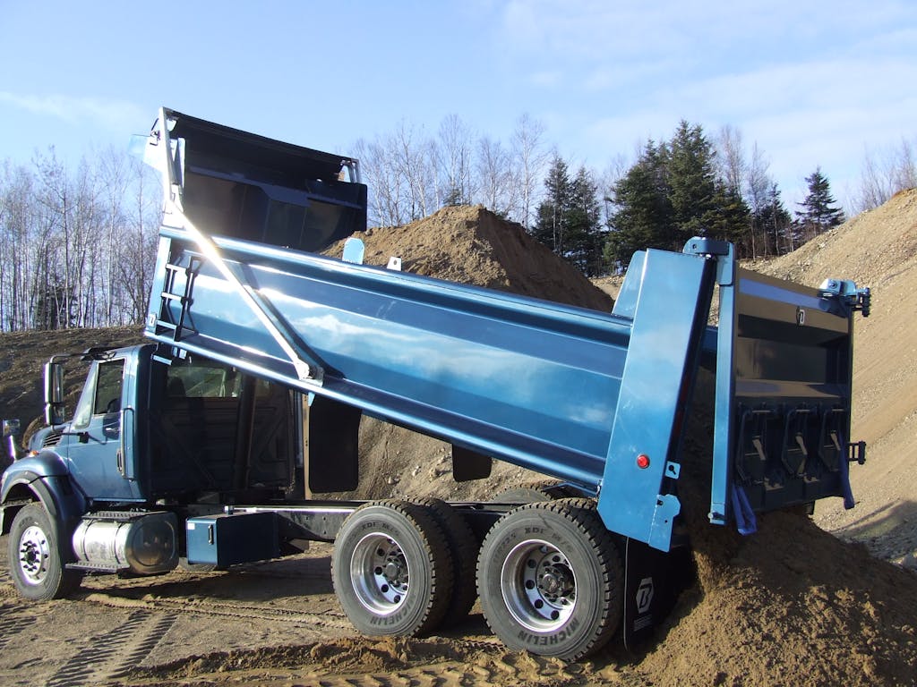 A blue truck lifts its dump body into the air to unload a delivery of dirt. 